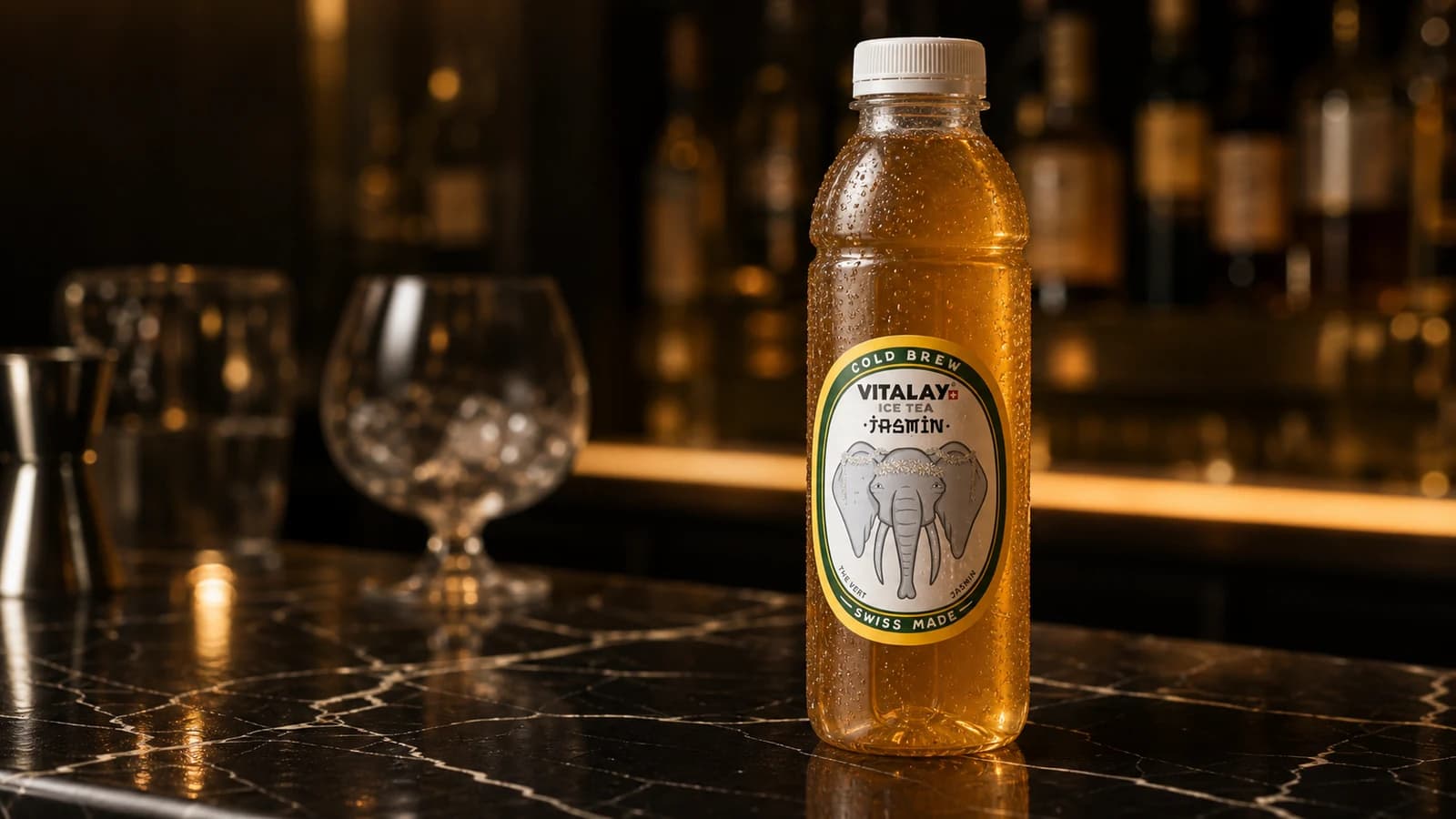 Bouteille Vitalay Thé froid Jasmin posée sur bar d'hôtel en marbre sombre, verre à eau en fond flou, lumière basse de bar