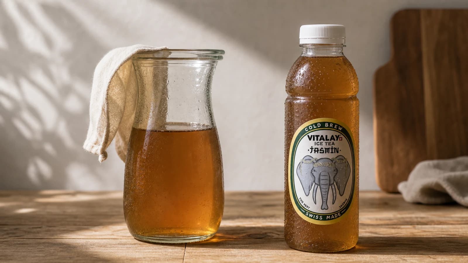 Carafe en verre avec thé glacé maison à gauche, bouteille Vitalay Thé froid Jasmin à droite, surface bois clair, lumière naturelle diffuse