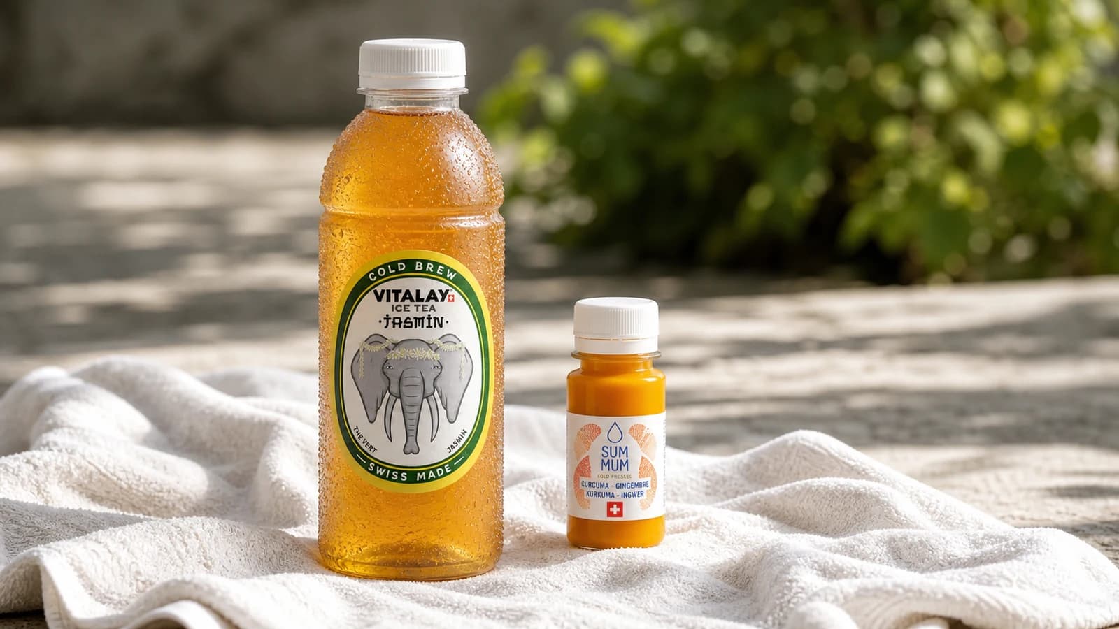 Bouteille Vitalay Thé froid Jasmin et shot SUM MUM gingembre posés sur serviette de sport, lumière naturelle extérieure