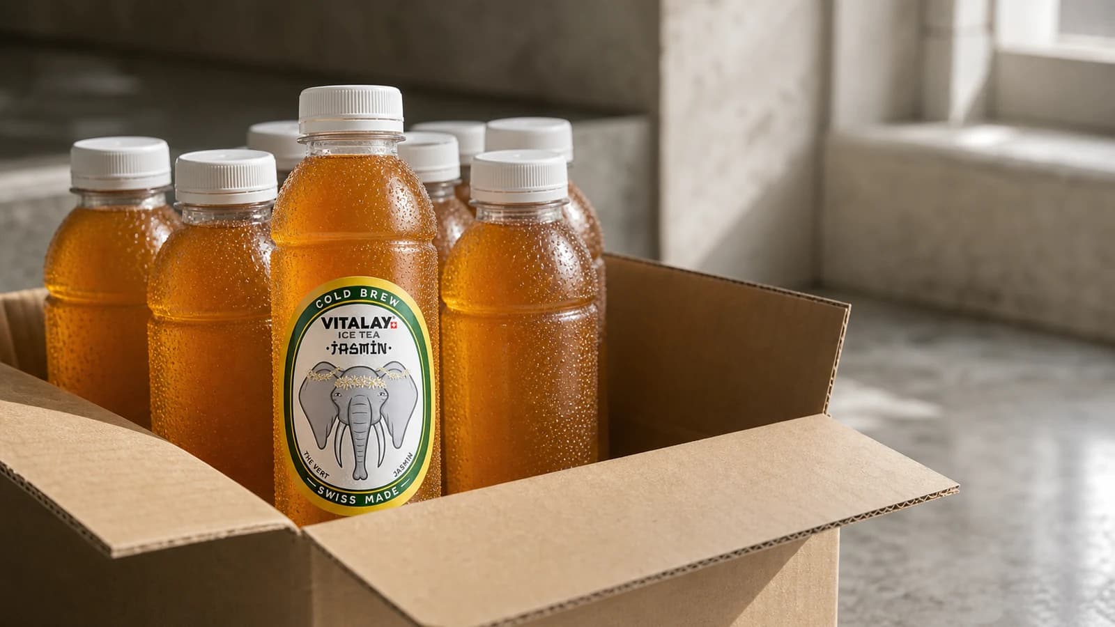 Carton Vitalay ouvert avec bouteilles Thé froid alignées, surface entrepôt propre, lumière naturelle latérale