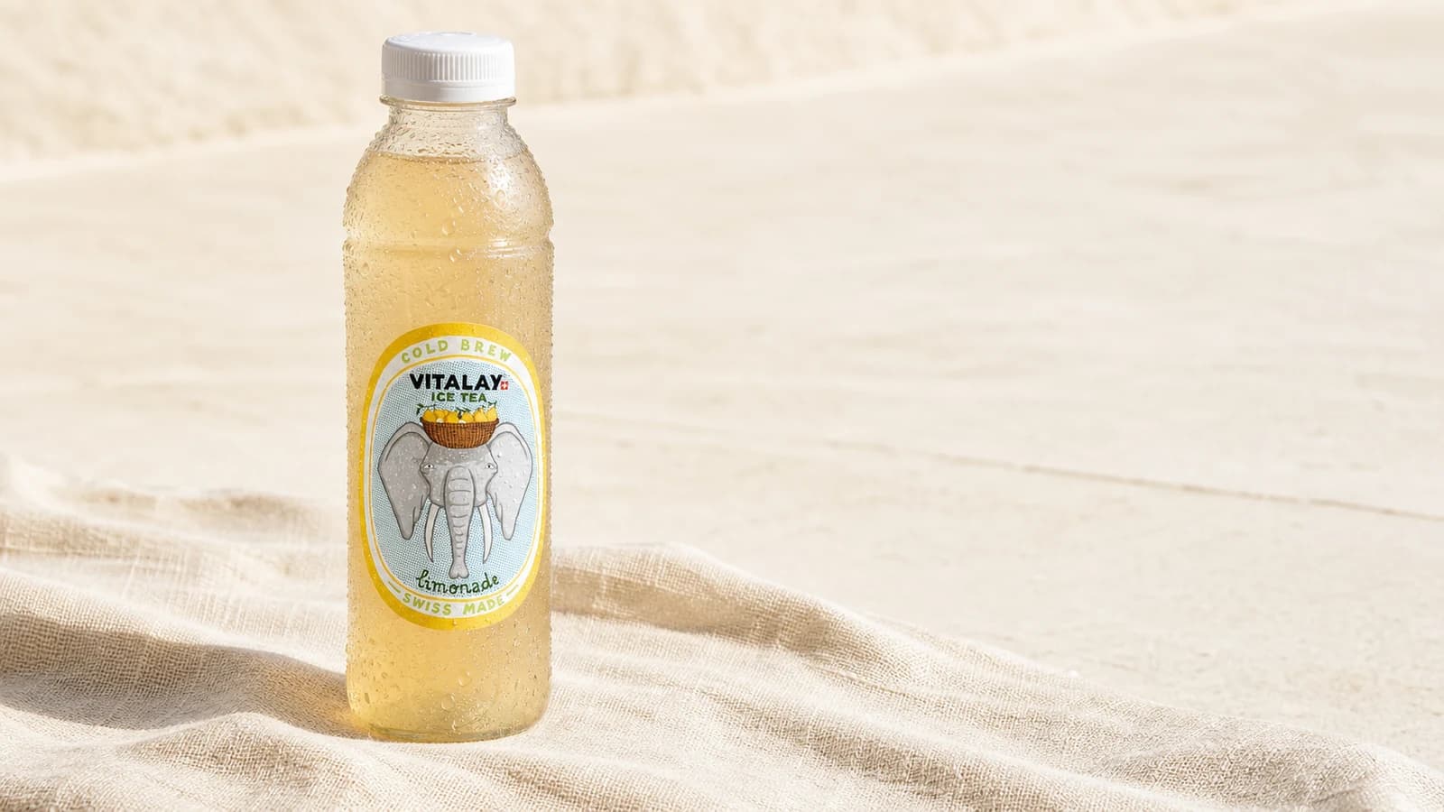 Bouteille Vitalay Thé froid Citron posée sur serviette de plage en lin, lumière d'été directe, fond sable